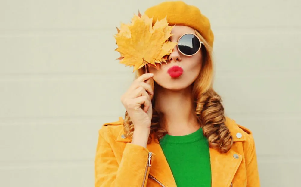 Autumn portrait of beautiful young woman covering her face with yellow maple leaves blowing her lips with red lipstick wearing a french beret on gray background