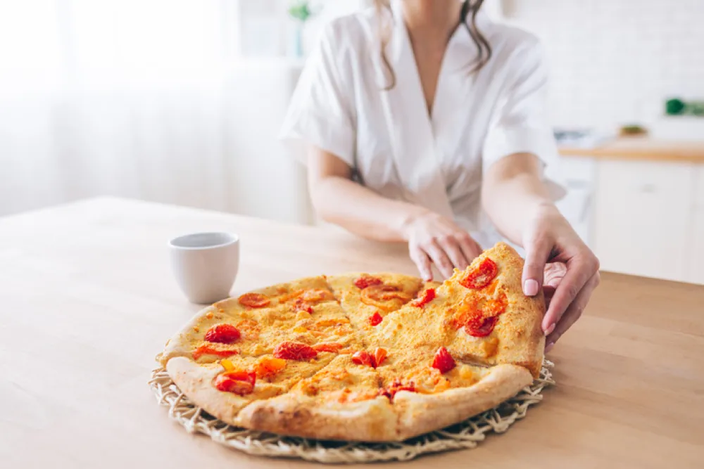 Cut view of woman in white dressing gown in kitchen. Taking piece of pizza. Small slice. Young housekeeper live carefree life